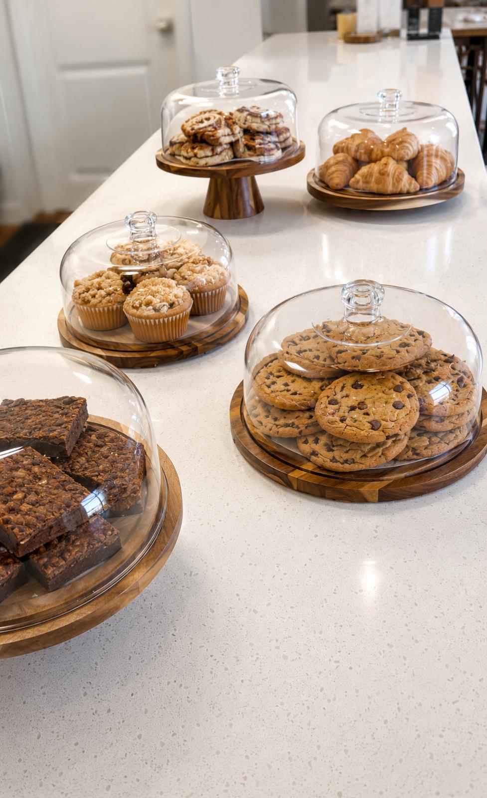 Wooden cake stands under glass domes holding chocolate chip cookies, brownies, muffins and croissants.