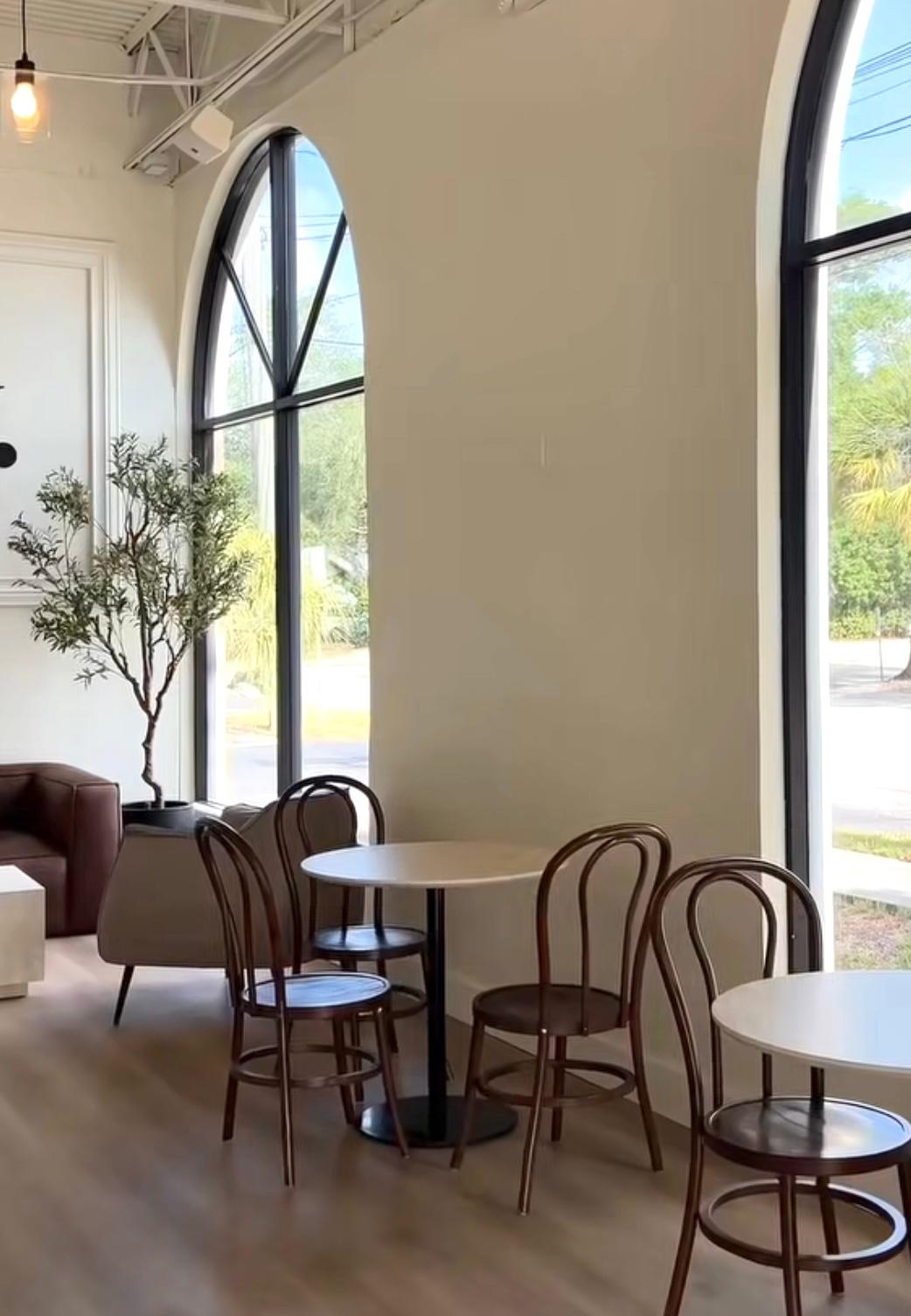 Two soaring arched windows pour daylight over a marble café table and an olive tree.